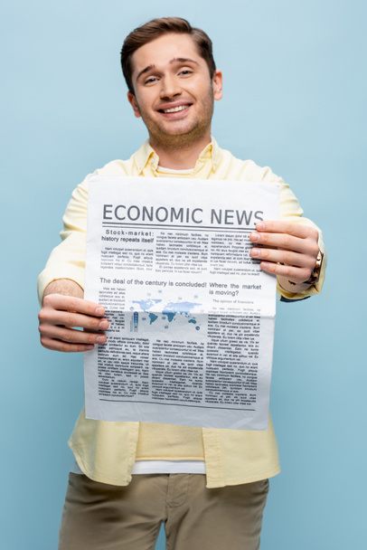 joven feliz en camisa sosteniendo periódico económico aislado en azul - Foto, Imagen