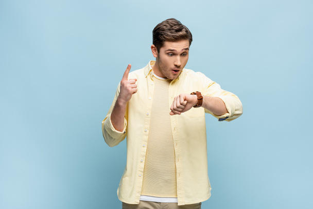 jonge man in geel shirt kijken naar polshorloge en wijzen met de vinger op blauw - Foto, afbeelding