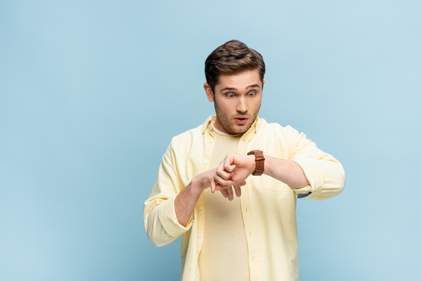 hombre joven sorprendido en camisa amarilla mirando reloj de pulsera en azul - Foto, Imagen