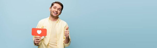 Kırmızı kağıt kalp tutan gömlekli mutlu bir adam ve mavi pankartta izole bir şekilde baş parmağını gösteriyor. - Fotoğraf, Görsel