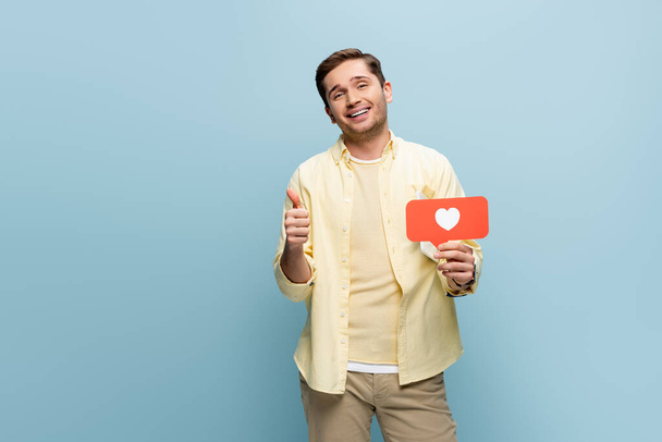 homme heureux en chemise tenant coeur en papier rouge et montrant pouce isolé sur bleu - Photo, image