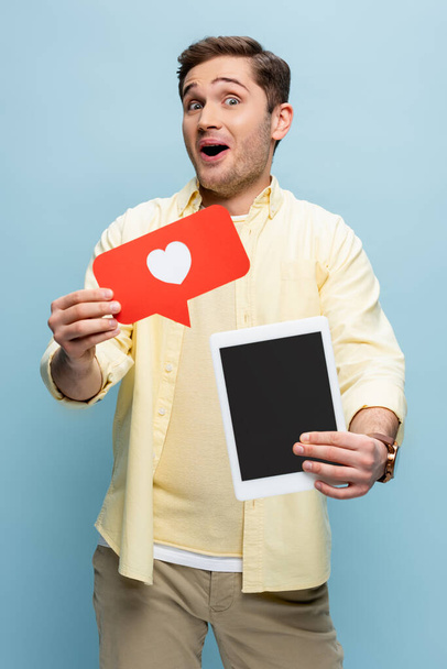 joven sorprendido en camisa sosteniendo corazón de papel y tableta digital con pantalla en blanco aislado en azul - Foto, Imagen