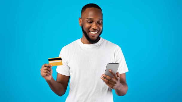 Χαρούμενος μαύρος τύπος που χρησιμοποιεί τηλέφωνο και πιστωτική κάρτα, μπλε φόντο - Φωτογραφία, εικόνα