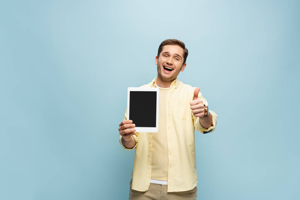 erstaunt Mann in gelbem Hemd hält digitales Tablet mit leerem Bildschirm und zeigt Daumen nach oben isoliert auf blau - Foto, Bild