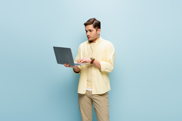 mladý muž ve žluté košili pomocí notebooku izolované na modré - Fotografie, Obrázek