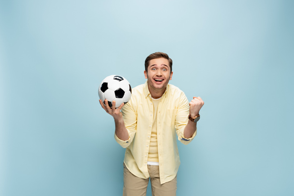счастливый человек в желтой рубашке, держащий в руках футбол и ликующий на голубом - Фото, изображение