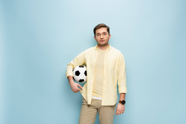 νεαρός άνδρας με κίτρινο πουκάμισο κρατώντας το ποδόσφαιρο στο μπλε - Φωτογραφία, εικόνα