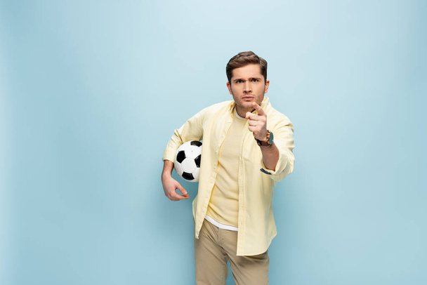 Mann im gelben Hemd hält Fußball in der Hand und zeigt mit dem Finger auf Kamera auf blau - Foto, Bild