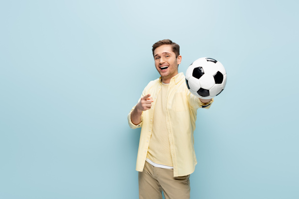 gelukkig man in geel shirt wijzend met vinger en houden voetbal op blauw - Foto, afbeelding