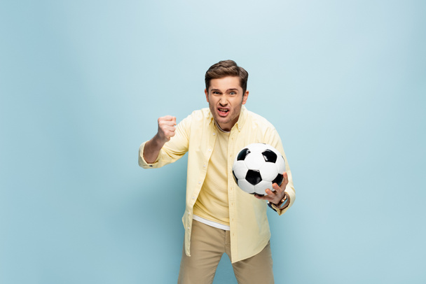 podrażniony mężczyzna w żółtej koszuli trzyma piłkę nożną na niebiesko - Zdjęcie, obraz
