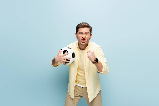 στρεσαρισμένος άντρας με κίτρινη μπλούζα κρατώντας ποδόσφαιρο στο μπλε - Φωτογραφία, εικόνα