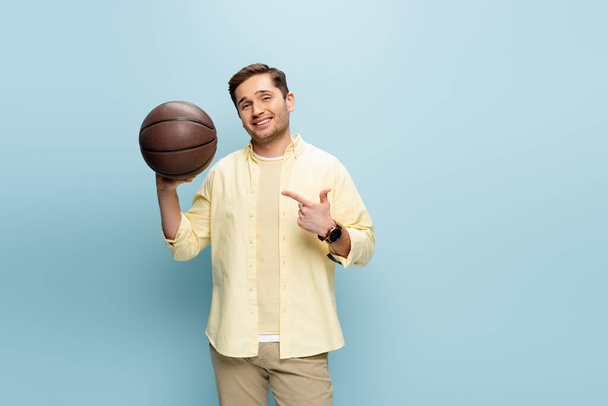 Ευτυχισμένος άντρας με κίτρινη μπλούζα που δείχνει με το δάχτυλο το μπάσκετ στο μπλε - Φωτογραφία, εικόνα