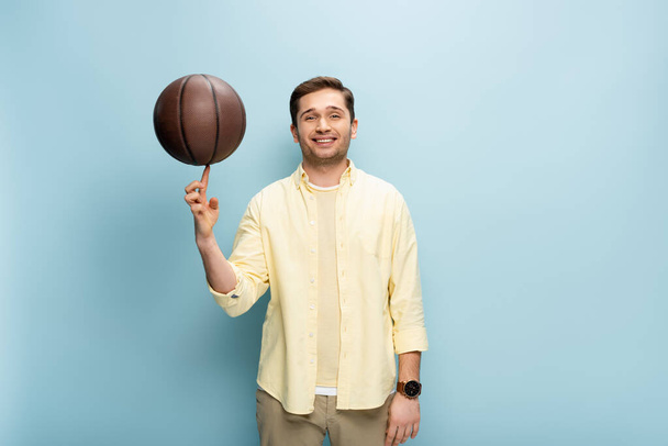 gelukkig man in geel shirt spinnen basketbal op vinger op blauw - Foto, afbeelding