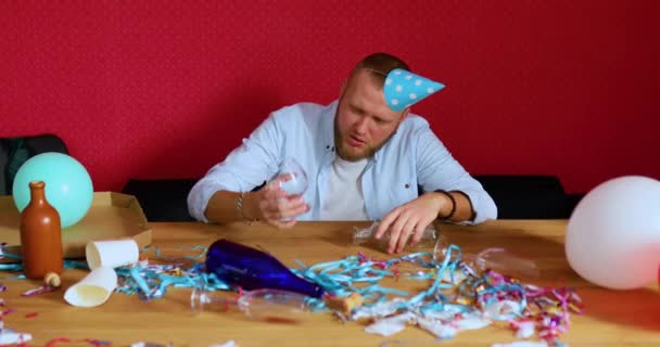 Hombre barbudo durmiendo en la mesa en la habitación desordenada en gorra azul después de la despedida de soltero o cumpleaños, sostenga una copa de brandy, hombre cansado después de la fiesta en casa - Metraje, vídeo