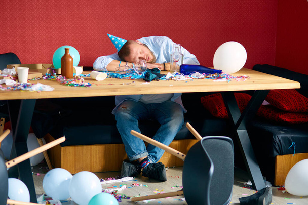 Mies nukkuu pöydässä sininen korkki sotkuisessa huoneessa syntymäpäiväjuhlien jälkeen, väsynyt mies juhlien jälkeen kotona - Valokuva, kuva
