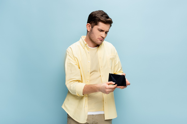 triste giovane uomo in camicia gialla guardando portafoglio vuoto su blu - Foto, immagini