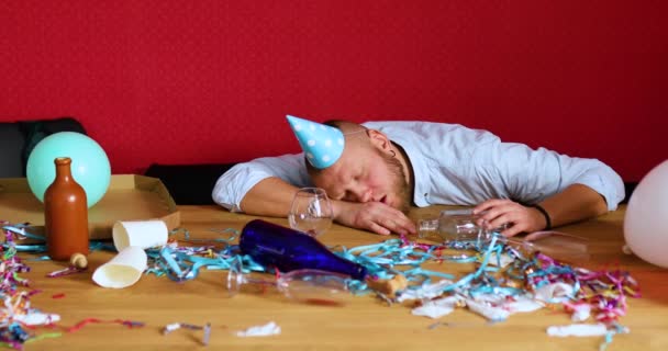 Baard man slapen aan tafel in rommelige kamer in blauwe pet na vrijgezel of verjaardagsfeestje, houd een glas cognac, vermoeide man na feestje thuis - Video