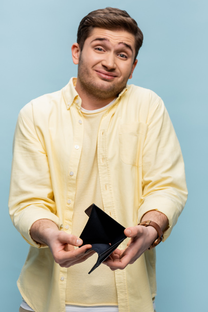 недовольный мужчина в желтой рубашке держит черный пустой бумажник изолирован на синий - Фото, изображение