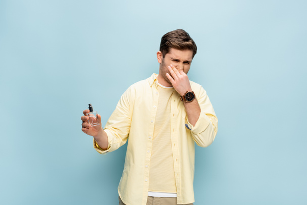walgelijke man in geel shirt bedekken neus en houden fles met parfum op blauw - Foto, afbeelding