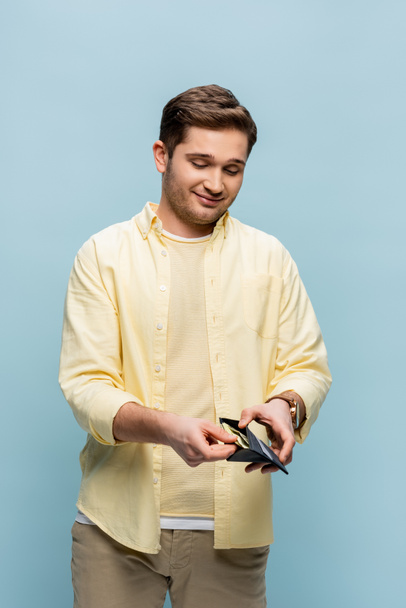 gelukkig jongeman in geel shirt houden portemonnee met dollars geïsoleerd op blauw - Foto, afbeelding