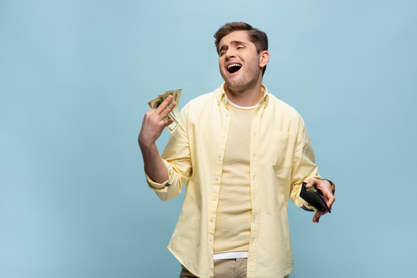 богатый молодой человек в желтой рубашке с бумажником и размахивая долларами изолированы на синий - Фото, изображение