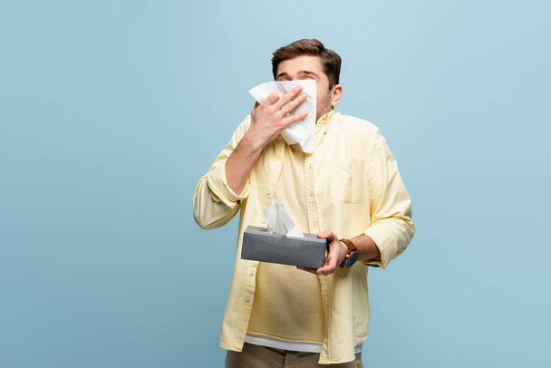 man die de mond bedekt met een servet terwijl hij niest en de weefseldoos geïsoleerd houdt op blauw - Foto, afbeelding