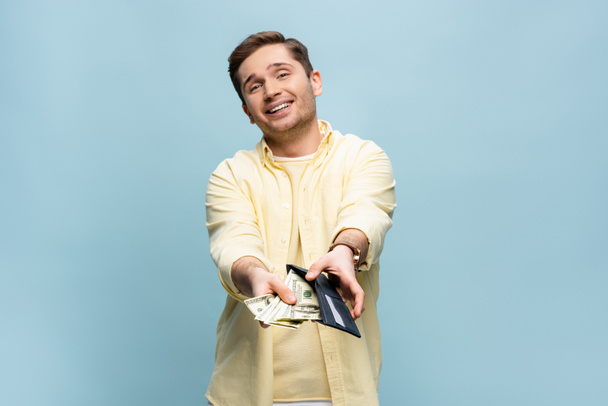 hombre feliz con camisa amarilla sosteniendo billetera con dólares aislados en azul - Foto, imagen