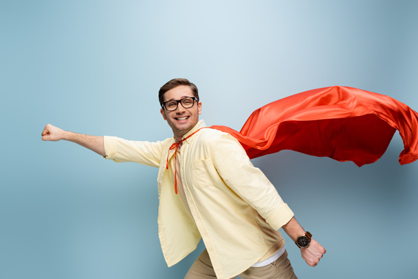 szczęśliwy superbohater w okularach i czerwonej pelerynie stojącej z wyciągniętą dłonią na niebieskim - Zdjęcie, obraz