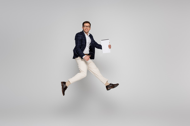 full length of cheerful businessman in glasses holding paper while jumping on grey - Photo, Image