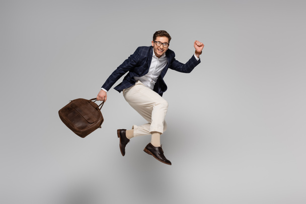 longitud completa del alegre hombre de negocios sosteniendo bolsa de cuero y levitando sobre gris - Foto, Imagen