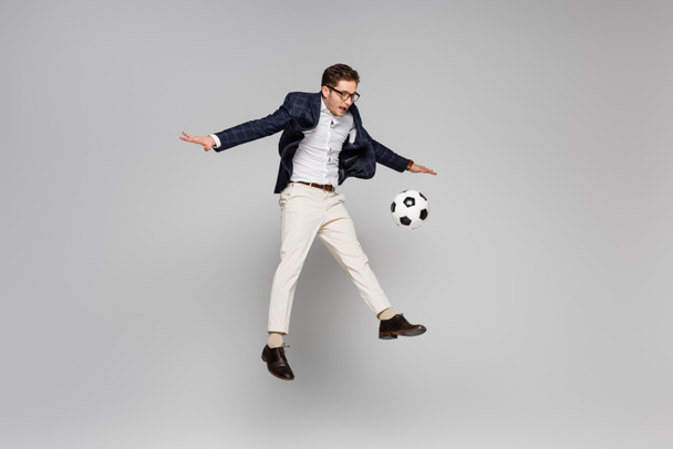 full length of cheerful businessman playing football while levitating on grey - Photo, Image