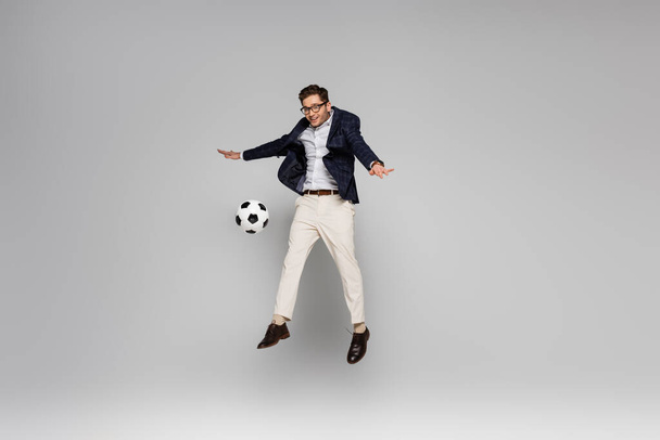 полный рост улыбающийся бизнесмен, играющий в футбол, пока левитирует на сером - Фото, изображение
