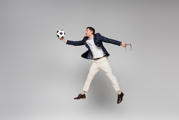 longitud completa de hombre de negocios sonriente sosteniendo gafas y jugando al fútbol mientras levita en gris - Foto, Imagen