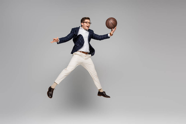 longitud completa de hombre alegre en gafas jugando baloncesto y levitando sobre gris - Foto, imagen