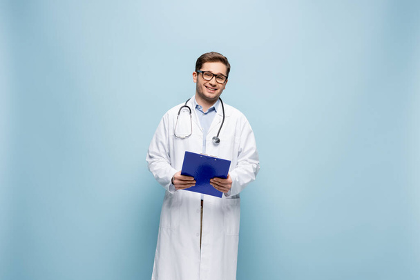 positiivinen lääkäri valkoisessa takissa pitäen leikepöytää ja katsoen kameraa sinisellä - Valokuva, kuva