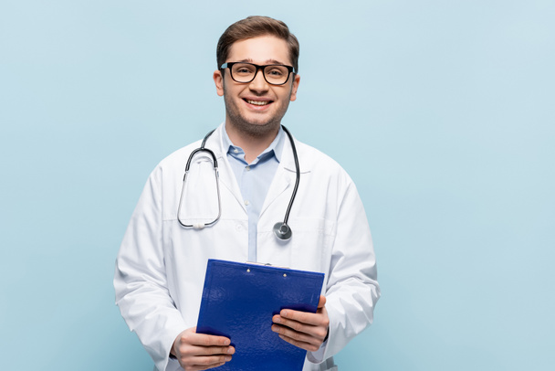 pozytywny lekarz w okularach i białym płaszczu trzymając schowek i patrząc na aparat odizolowany na niebiesko - Zdjęcie, obraz