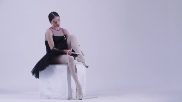 Junge Ballerina im schwarzen Kleid zieht Spitzenschuhe im weißen Studio an - Filmmaterial, Video