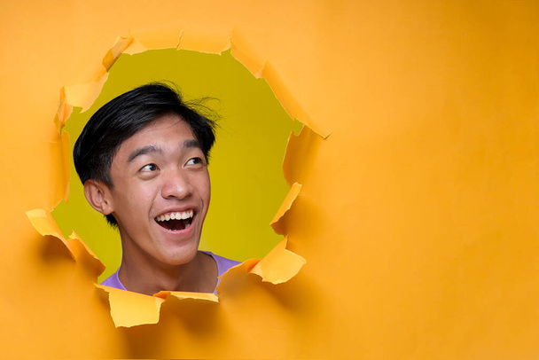 Heureux jeune homme asiatique souriant regarder un espace de copie, pose à travers trou de papier jaune déchiré. Publicité Concept - Photo, image