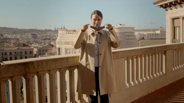 Giovane donna europea in piedi da sola sul balcone in un trench beige si sta mettendo una maschera viso. Ritratto frontale medio full shot immagine ad alta risoluzione - Foto, immagini