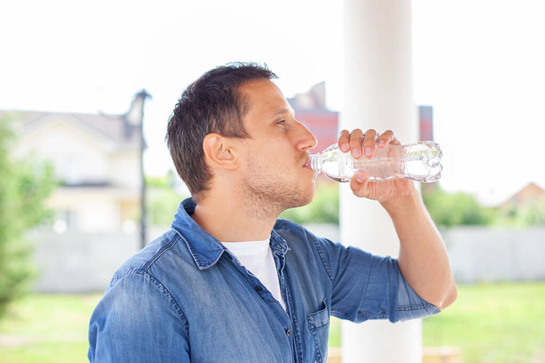 Primer plano de un hombre bebiendo agua de la botella al aire libre. Un hombre refrescándose. Hombre sediento bebiendo agua de una botella de plástico en el parque de verano. Concepto de salud. Hombre joven bebiendo agua después del deporte - Foto, Imagen