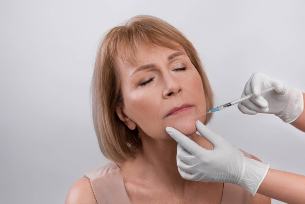 Esthetische kosmetologie. Senior vrouw krijgt hyaluronzuur of collageen injectie op lichte studio achtergrond - Foto, afbeelding