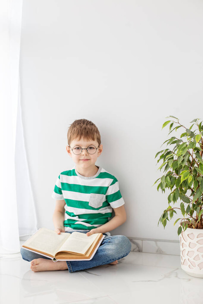 Un niño con una camiseta de rayas verdes se sienta en el suelo leyendo con entusiasmo un libro, pasa su tiempo libre estudiando, junto a un árbol verde. - Foto, imagen