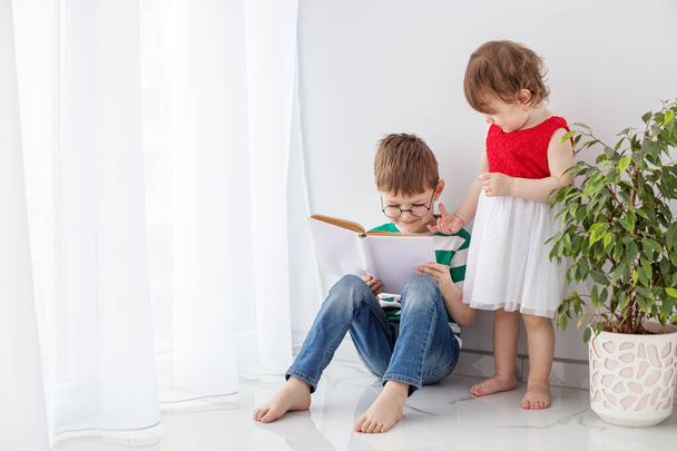 Маленький хлопчик і дівчинка проводять дозвілля разом, читаючи книгу, у світлій кімнаті поруч з домашнім рослиною
. - Фото, зображення