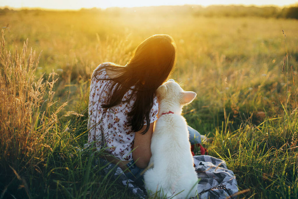 Happy woman with cute white puppy sitting in summer meadow and looking at warm sunset light. Casual young female cuddling with adorable swiss shepherd  puppy. Happiness. Vacation with pet - Photo, Image