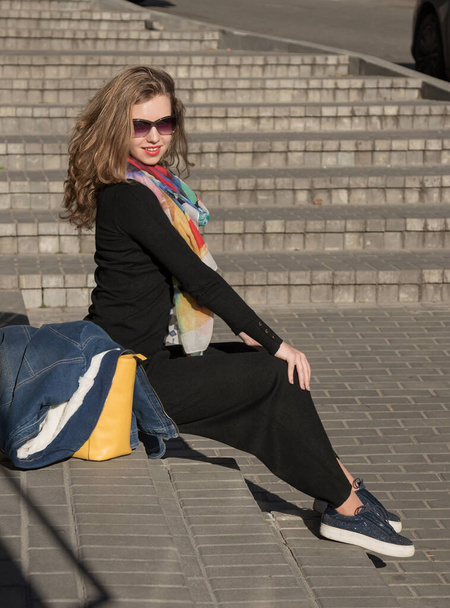 κομψό κορίτσι κάθεται στα σκαλιά με γυαλιά ηλίου και ένα σκούρο κοστούμι. Λεπτομέρειες για τα γυναικεία street-style ρούχα. - Φωτογραφία, εικόνα