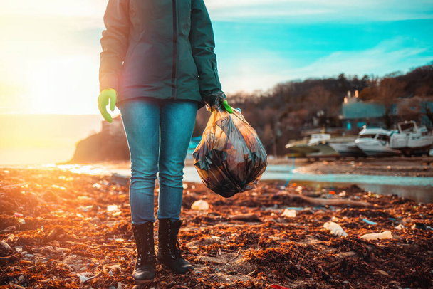 Día de la Tierra. Una voluntaria está en una playa fangosa con una bolsa de basura. Puesta de sol. - Foto, Imagen