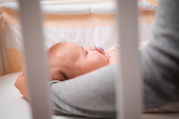 Ένα μωρό είναι ξαπλωμένο στην κούνια με μια πιπίλα, η μητέρα χαϊδεύει το κεφάλι του. Πλάγια άποψη, μέσα από τα κάγκελα της κούνιας. - Φωτογραφία, εικόνα