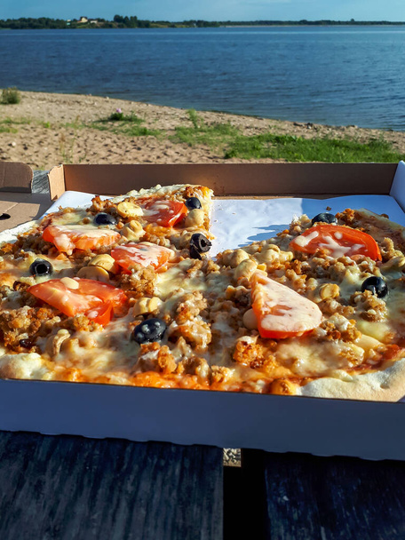 砂と水の近くの木製のテーブルの上に箱の中に混合肉、トマト、オリーブとチーズとピザ屋外 - 写真・画像