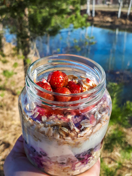 Zdrowe śniadanie na noc owies z truskawkami, jagodami i jogurtem w szklanym słoiku z pięknym tłem na świeżym powietrzu - drzewa, trawa i woda. Koncepcja przygotowywania żywności i przygotowywania posiłków - Zdjęcie, obraz