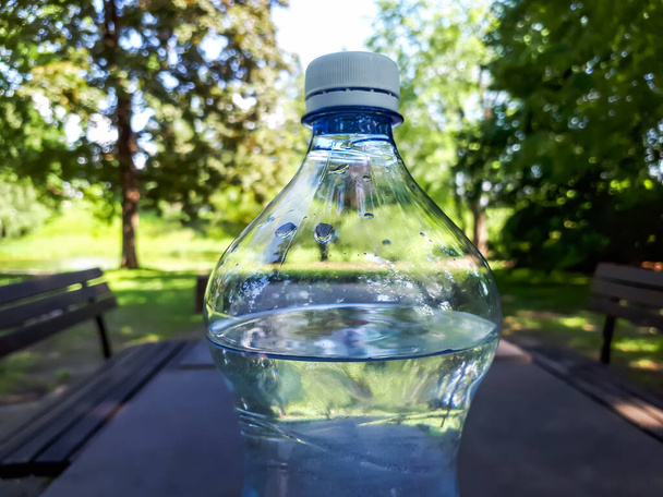 Primo piano colpo di bottiglia di plastica con tappo bianco riempito con acqua sul tavolo da campeggio all'aperto marrone con natura verde alberi, erba e sfondo. Importanza dell'idratazione e dell'acqua potabile - Foto, immagini
